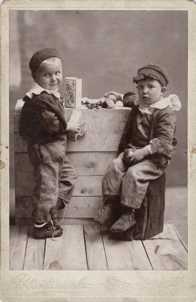 Twins wearing hats, jackets, and pants are sitting and standing near a wooden box with food displayed on it.
