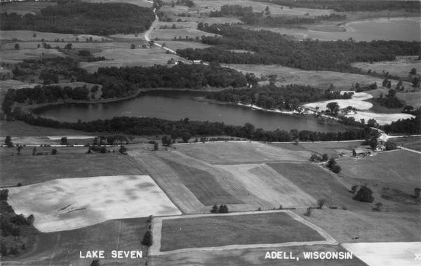 Aerial view of Lake Seven, surrounded by trees. Agricultural fields fill the foreground. Another lake is in the upper right. Caption reads: "Lake Seven Adell, Wisconsin".