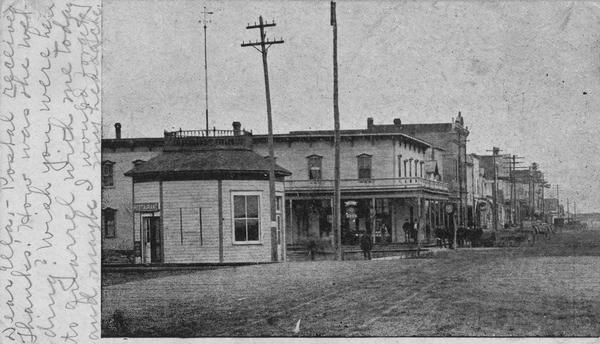 View down Keller Avenue towards businesses along the left. A round or octagon shaped building on the far left has a sign that reads: "Restaurant". Further down on the left people are standing near a large building with a porch, and a balcony above. Horse-drawn vehicles are along the curb.