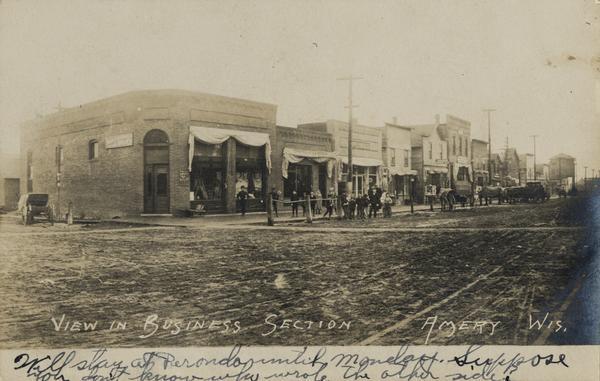 Business section of downtown. View across unpaved road towards business along the left. A group of children and adults  are posing standing in the road near the curb. Caption reads: "View in Business Section, Amery, Wisconsin." 