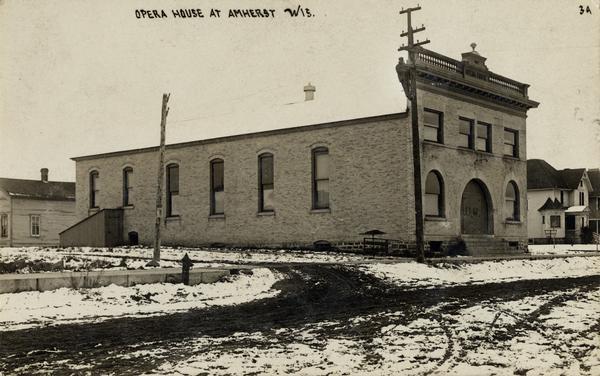View across unpaved road towards the Opera House, with a man high up on a telephone pole in front of the building. Snow is on the ground. Caption reads: "Opera House at Amherst, Wis."