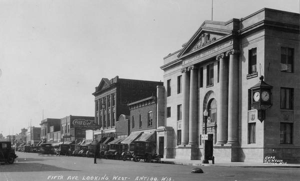 Caption reads: "5th Ave. Looking West — Antigo, Wis." View across intersection towards the Langlade National Bank on the right corner, which has a clock on the corner of the building. Automobiles are parked at an angle along both sides of the street. 