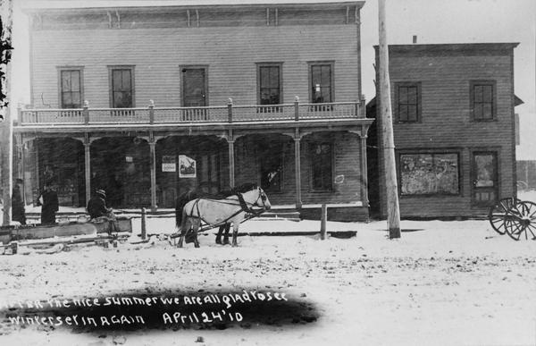 The Hoo Hoo Club Hotel, a lumbermen's social organization. Two men are standing near a man driving a sled carrying milk cans pulled by two horses in front of the building. It is snowing and snow is on the ground. Caption reads: "After the nice summer we are all glad to see winter set in again. April 24 '10."