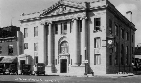 View across street towards the Langlade National Bank on the corner. A clock is on the side of the building, and automobiles are parked at an angle along the curb.