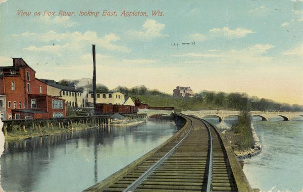 View of the Fox River, looking east, from railroad tracks. There is a bridge further down on the right. Buildings line the shoreline on the left, and a large building is on a hill in the distance. Caption reads: "View on Fox River, looking East, Appleton, Wis."