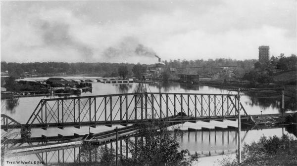 Elevated view of a railway bridge over the river, with industrial buildings along the shoreline in the background.