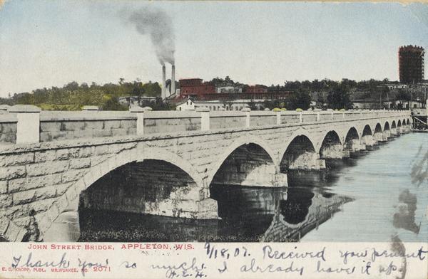 John Street Bridge crossing the river on the left, with industrial buildings and smokestacks in the distance on the opposite shoreline. Caption reads: "John Street Bridge, Appleton, Wis."