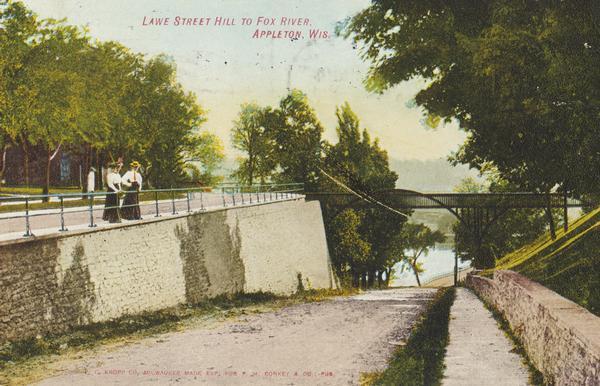 Scenic view of two women standing along a railing on a sidewalk above a stone wall viewing the way to the river. Caption reads: "Lawe Street Hill to Fox River, Appleton, Wis."