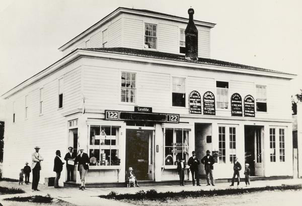 Text reads: "Masonic Hall at the corner of College Avenue and Oneida Street; built in 1855 by Theodore Conkey, it burned in 1874. The Zuehlke Building was built on this site later.  The Clegett Barber Shop was at the extreme right. The Masonic rooms were on the top story, and various shops and businesses below. The two boys on Oneida Street are Fred and Frank Jerrard. The tall man is Dyer Fox; next is Mr. Burdick. W.F. Montgomery stands in the doorway, newspaper in hand. The small boy with the dog is Beverly Gilmore, and the dog belonged to Mark Lyon. Prof. Cuthbert, music teacher, wears striped pantaloons."