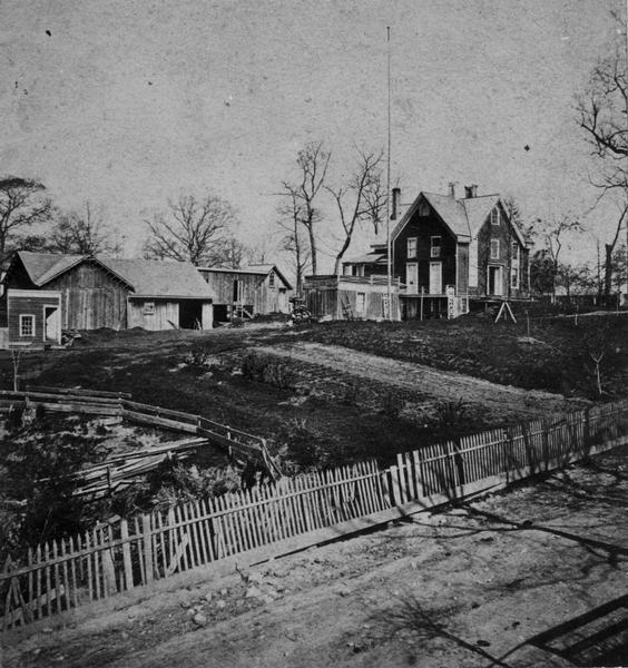 Elevated view across road and fence towards two houses and a yard, bordered by a damaged, white picket fence. The house burned down in 1867.