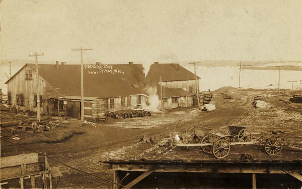 Exterior view of a machine shop, with a wagon in the foreground. A body of water is behind the shop, and railroad tracks curve around to the right. A sign near the tracks reads: "Railroad Crossing". Caption reads: "Machine Shop, Arbor Vitae, Wis."