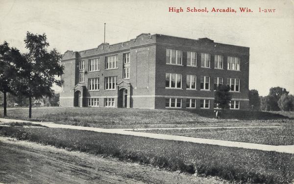 Exterior view of the two-story brick High School building with basement. A bare flagpole is on the roof. Caption reads: "High School, Arcadia, wis."