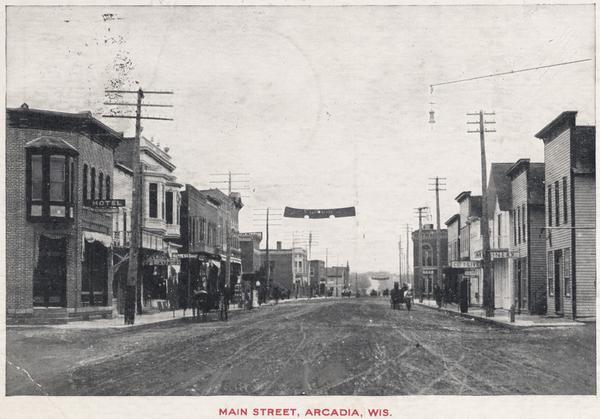 View down Main Street, with a hotel on the left side and horse-drawn wagons on the unpaved street. Pedestrians are on the sidewalks. Caption reads: "Main Street, Arcadia Wis."