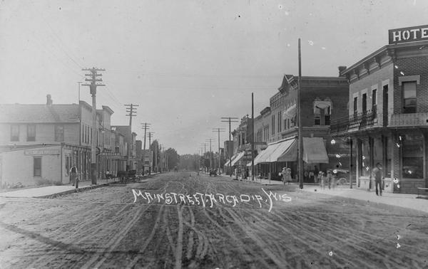Unpaved Main Street with many storefronts and a hotel on the right hand side. Pedestrians are on the sidewalks. Caption reads: "Main Street, Arcadia, Wis".