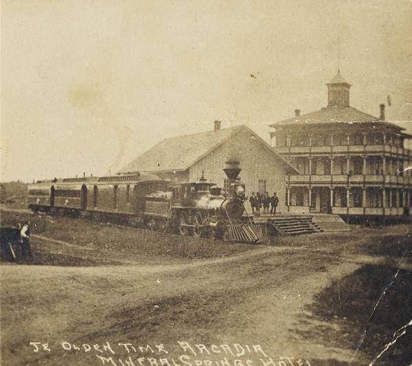 View across unpaved road towards a group of men standing on the platform at the train depot, with the Mineral Springs Hotel behind and to the right. Another man is standing across the road on the left. A locomotive with four railroad cars is paused on the tracks. The hotel has three stories with wraparound porches and balconies. Caption reads: "Ye Olden Time Arcadia Mineral Springs Hotel".