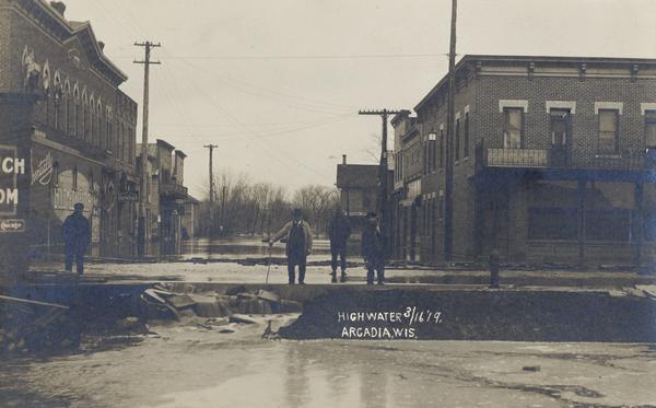 View showing the flood conditions on city streets from the 1919 Flood on the Trempealeau River. Four men are surveying the damage. Caption reads: "Highwater, Arcadia, Wis." 