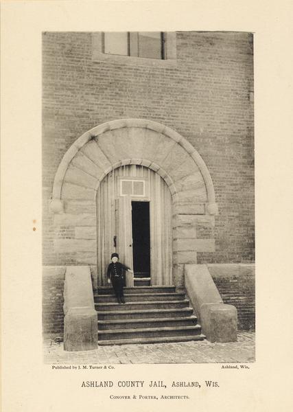 Text on front reads: "Ashland County Jail, Ashland, Wis." and "Conover & Porter, Architects." A young boy standing on the steps near the open door of the arched entrance to the Ashland County Jail. 