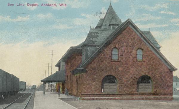 Caption reads: "Soo Line Depot, Ashland, Wis." It was built in 1890 of brownstone in the Richardson Romanesque style. Pedestrians stand on the platform of the depot. Railroad cars are on the second set of tracks on the left. It is listed in the State and National Register of Historic Places.<p>The railroad company was founded in 1888 and named for the Minneapolis, St. Paul and Sault Ste. Marie Railroad (MStP&SSM), which was commonly known as the Soo Line after the phonetic spelling of Sault.</p>