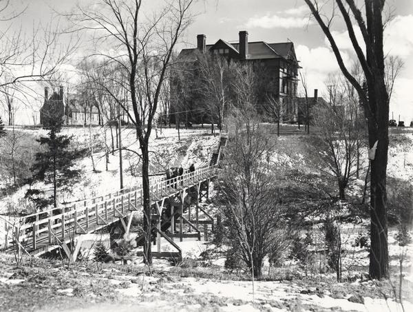 Wheeler Hall at Northland College, during winter. A group of people are on a bridge over the Bay City Creek, surrounded by trees, in the foreground. Wheeler Hall is a three story building with basement and attic. Built in 1893 of red brick and brownstone in the Romanesque Revival style. An extensive Property Record is on the Wisconsin Historical Society website, see "Property Record, 1411 Ellis Ave, Architecture and History Inventory." It is listed on the State and National Register of Historic Places.