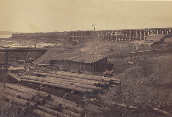 View of the ore dock of the Milwaukee, Lake Shore, and Western Railroad. Large logs, buildings and a railroad bridge are in the foreground.