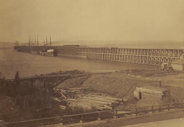 Ore dock of the Milwaukee, Lake Shore, and Western Railroad, with a pile of logs in the foreground and docked ships in the background.