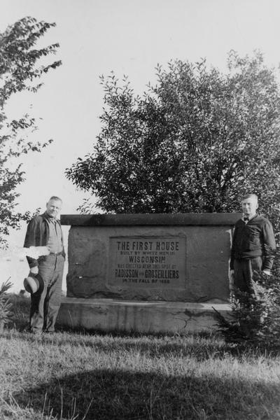 View of two men posing on either side of the Radisson and Groseilliers House historical marker. The house was the first built by white men in Wisconsin and was located in the vicinity of Ashland at the southern extremity of Chequamegon Bay.