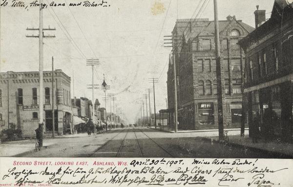 Text on front reads: "Second Street, Looking East, Ashland, Wis." An unpaved street with two sets of streetcar tracks in the center. Buildings, storefronts, sidewalks and pedestrians line the street.