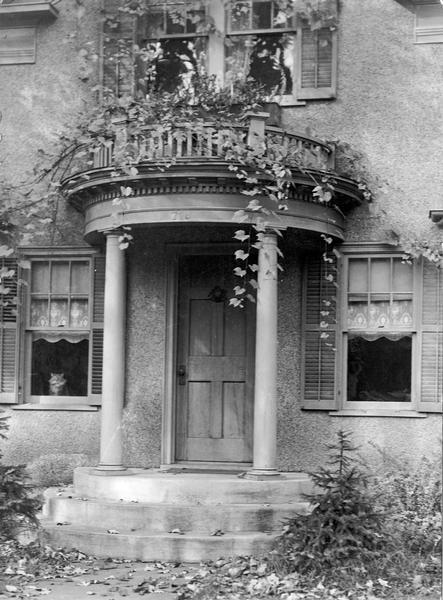 Exterior of the residence of Edwin B. and Alice Kent Trimpey, showing front entrance with columns, with a porch and plants above. A cat looks out of the window on the left. The home was located at 719 Oak Street.