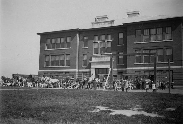 View of Ondossagon, a consolidated school. View across lawn towards a large group of people standing near horses in front of the building.