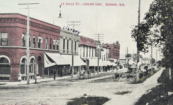 View of La Salle Street, looking east. Handwritten above a building in the center is "Library". Horse-drawn vehicles are along the right, and a person is driving a horse-drawn vehicle up the street on the left.  Caption reads: "La Salle St., Looking East, Barron, Wis."