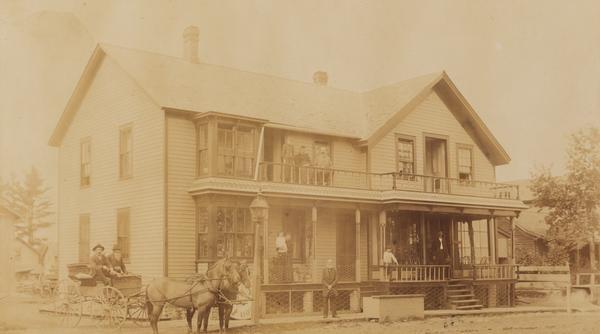 View of the Park House Hotel (called the Commercial House before 1888), which burned down in 1892. Two people are sitting in a horse-drawn vehicle on the left, and other people are posing on the porch and the balcony of the hotel.
