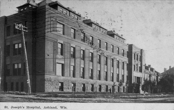 Caption reads: "St. Joseph's Hospital, Ashland, Wis." Front facade and grounds of the four-story, red brick hospital built in 1905. The basement level is built of stone. Two people are standing on the sidewalk. It was demolished in 1973.