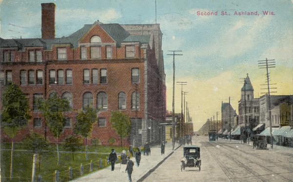 Slightly elevated view of Second Street, with an automobile on the street, two sets of streetcar tracks, pedestrians on the sidewalk, and a large brick building on the left. Caption reads: "Second St., Ashland, Wis."
