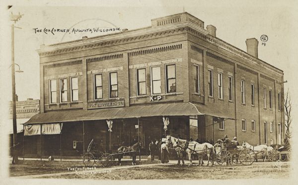 View from street of a group of people, including a number of horse-drawn vehicles, standing outside of the main entrance to the Cox Brothers company on Cox Corner.