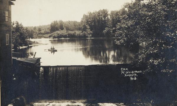 Front view of dam and waterfall at Dells Pond. There is a building on the left of the dam. People  are fishing from a boat on the pond.