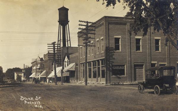 Photographic postcard of Stone Street. A car is parked in front of an auto repair store. Electric power lines run down the street, and a water tower is in the background. Caption reads: "Stone St., Augusta, Wis."