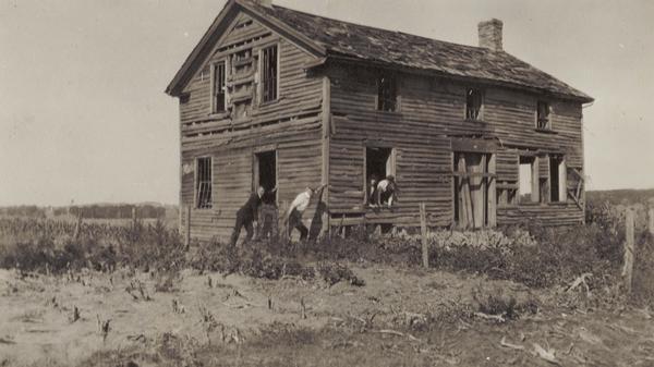 View of an old home or inn. Two people are looking out of an open window, and two men are standing outside leaning against the building.