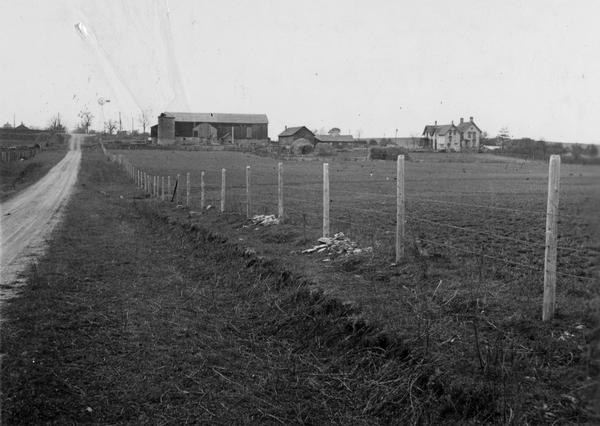 Distant view of Waterbury mansion, with field, road and fencing in the foreground.