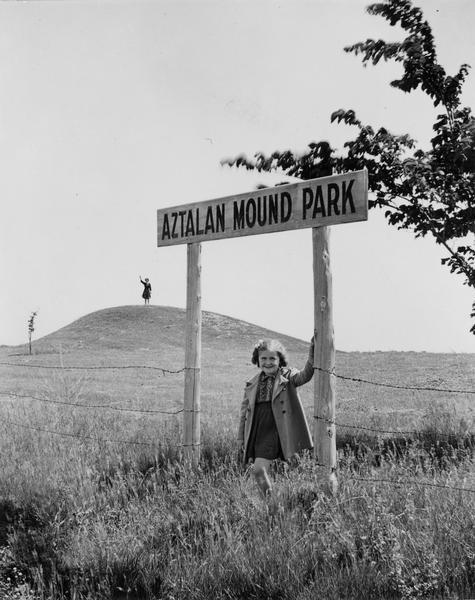 Little girl under the "Aztalan Mound Park" sign, in a field. Another person stands in the background on one of the mounds.