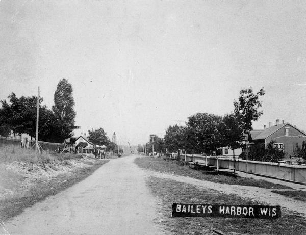 View down road. There is a long fence and a sidewalk on the right side. Houses and trees are on both sides of the road. Caption reads: "Baileys Harbor Wis".