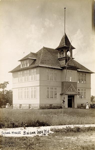Balsam Lake school house. A woman is standing on the right near the building. Caption reads: "School House Balsam Lake WIS".