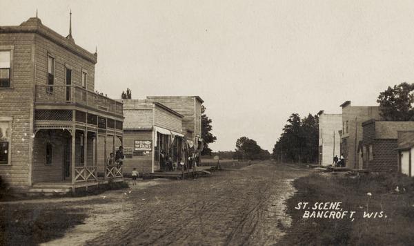 View down unpaved street with commercial buildings. Caption reads: "St. Scene, Bancroft, Wis."