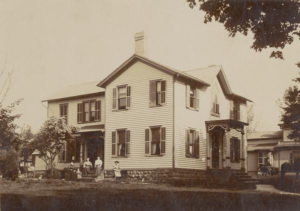 View across yard towards the Johnson family sitting on steps of their house at 110 Grove Street. A horse and buggy is parked in the front yard. Mrs. Johnson (left), T.S. Johnson (on cart). Dr. Johnson was pastor of the Assembly Presbyterian Church of Beaver Dam.