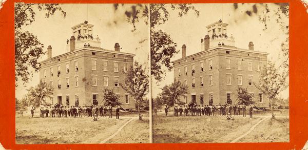 Stereograph view of the Middle College building. Groups of men are posing standing in front, and other men are posing at the entrance and in open windows. Another group of men are posing on the roof which has a widow's walk.