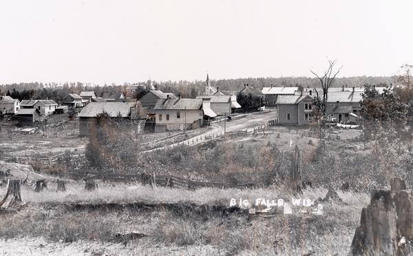 View from hill across field towards farmhouses, dwellings and churches along a road. In the foreground are stumps. Caption reads: "Big Falls, Wis."