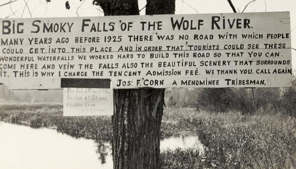 Sign for the toll road at Big Smoky Falls on the Wolf River. The sign reads: "Big Smoky Falls of the Wolf River. Many years ago before 1925 there was no road with which people could get into this place and in order that tourists could see these wonderful waterfalls we worked hard to build this road so that you can come here and veiw[sic] the falls also the beautiful scenery that surrounds it. This is why I charge the ten cent admission fee. We thank you, call again Jos. F. Corn A Menominee Tribesman."
