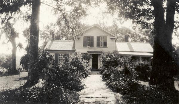 Front view of Man Mound House, with trees and a walkway which leads to the front door. The walkway appears to be on a mound.