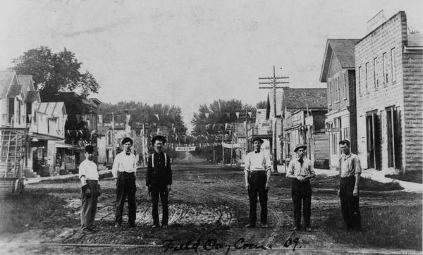 Group portrait of six men standing in the street. Behind them are storefronts in downtown Black Earth.