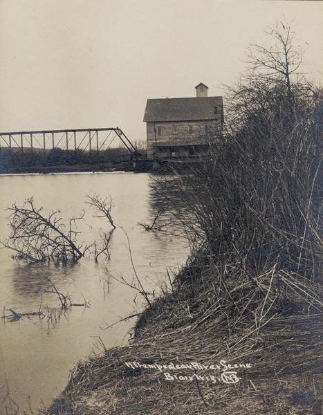 Trempealeau River and its banks, with a building and bridge in the background. Caption reads: "Trempealeau River Scene, Blair, Wis."