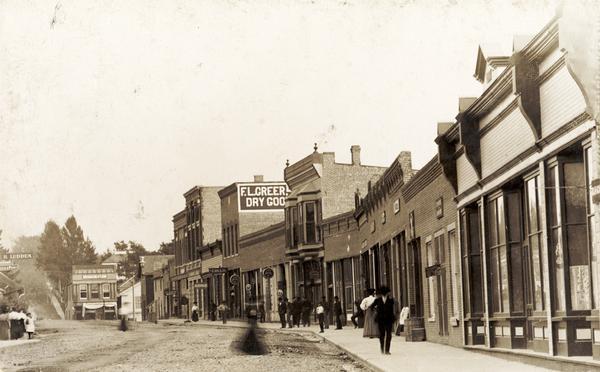 View down Main Street with pedestrians on the sidewalk on the right. A sign painted on the side of the building reads: "F.L. Greer Dry Goods".